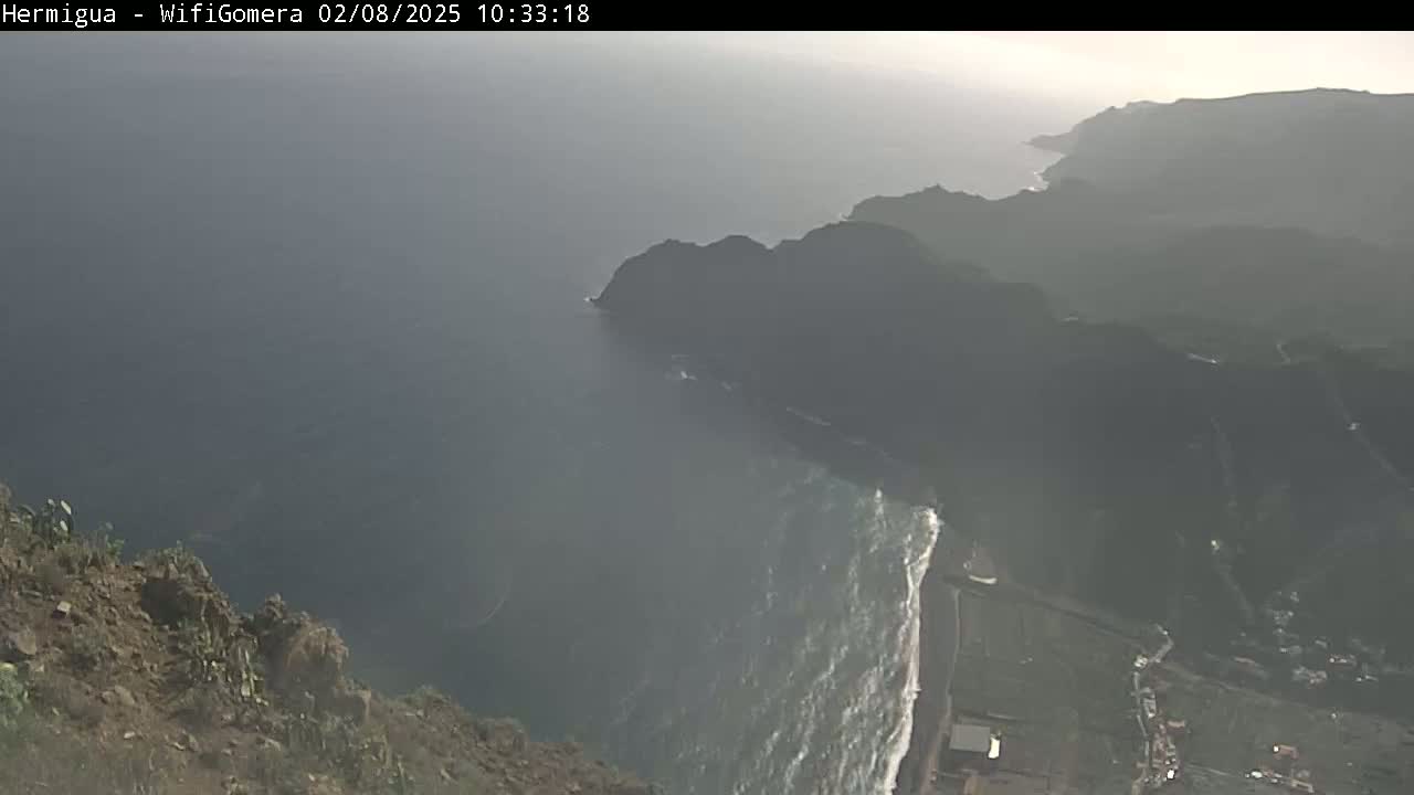 A hazy view from a clifftop shows a dark-colored coastline meeting a somewhat choppy ocean.
