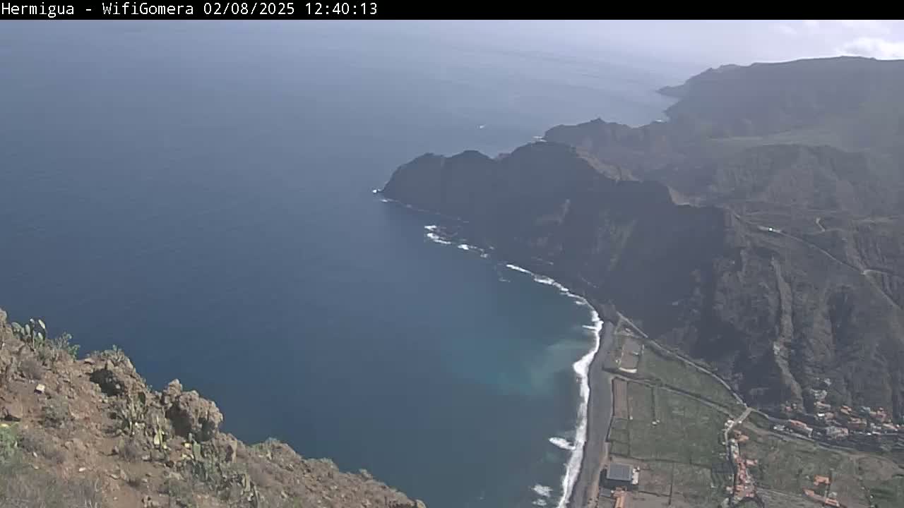 A high-angle view shows a rocky, mountainous coastline meeting a dark blue ocean under a clear sky.