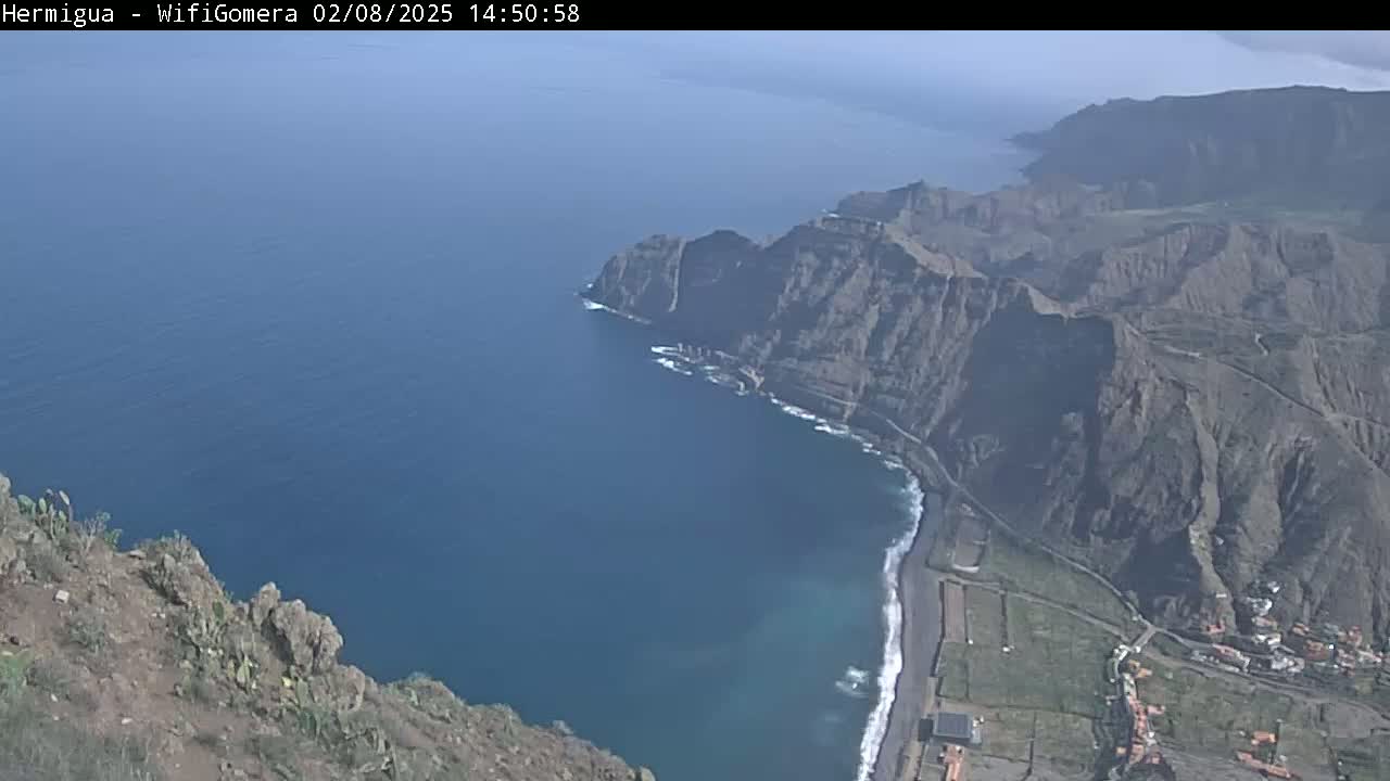 A high-angle view shows a rocky, mountainous coastline meeting a calm, dark-blue ocean under a clear sky.
