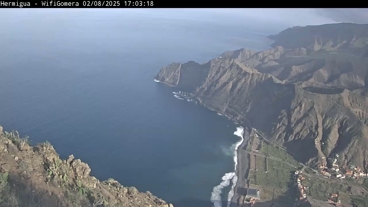 A high-angle view shows a rocky, mountainous coastline meeting a dark blue ocean under a mostly clear sky.
