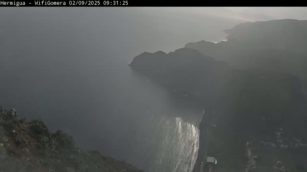 A hazy, overcast view shows a rocky coastline with a dark ocean meeting the land, possibly with some waves crashing against the shore.