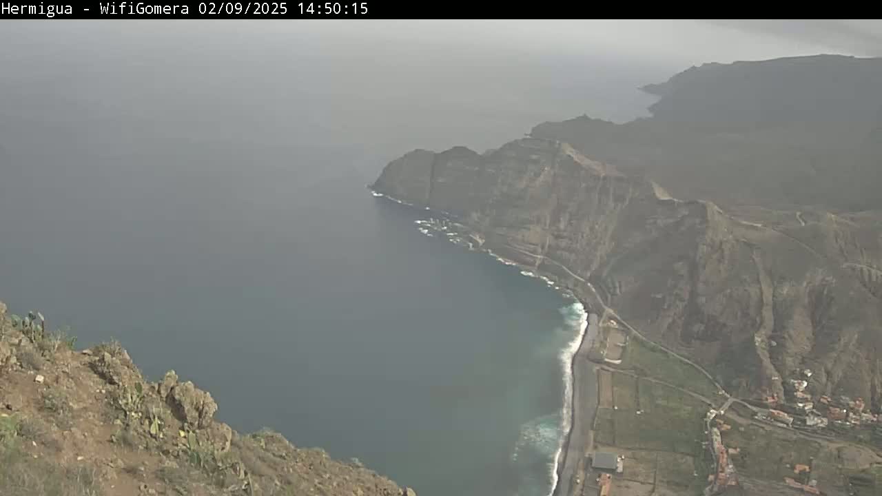 A hazy coastal view shows dark-colored cliffs meeting a dark-blue ocean with small waves breaking along a sandy beach at the base of the cliffs.