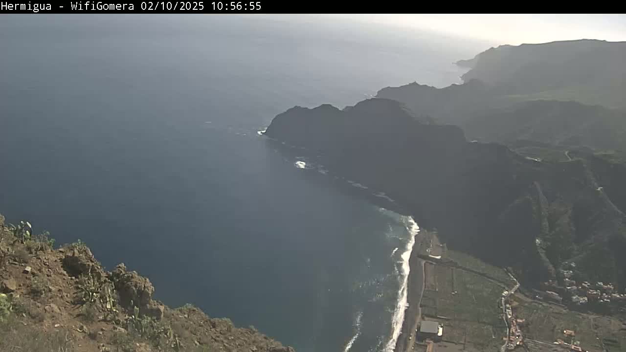 A hazy, high-angle view shows a dark-blue ocean meeting a rocky, mountainous coastline with a small beach and some flat land.