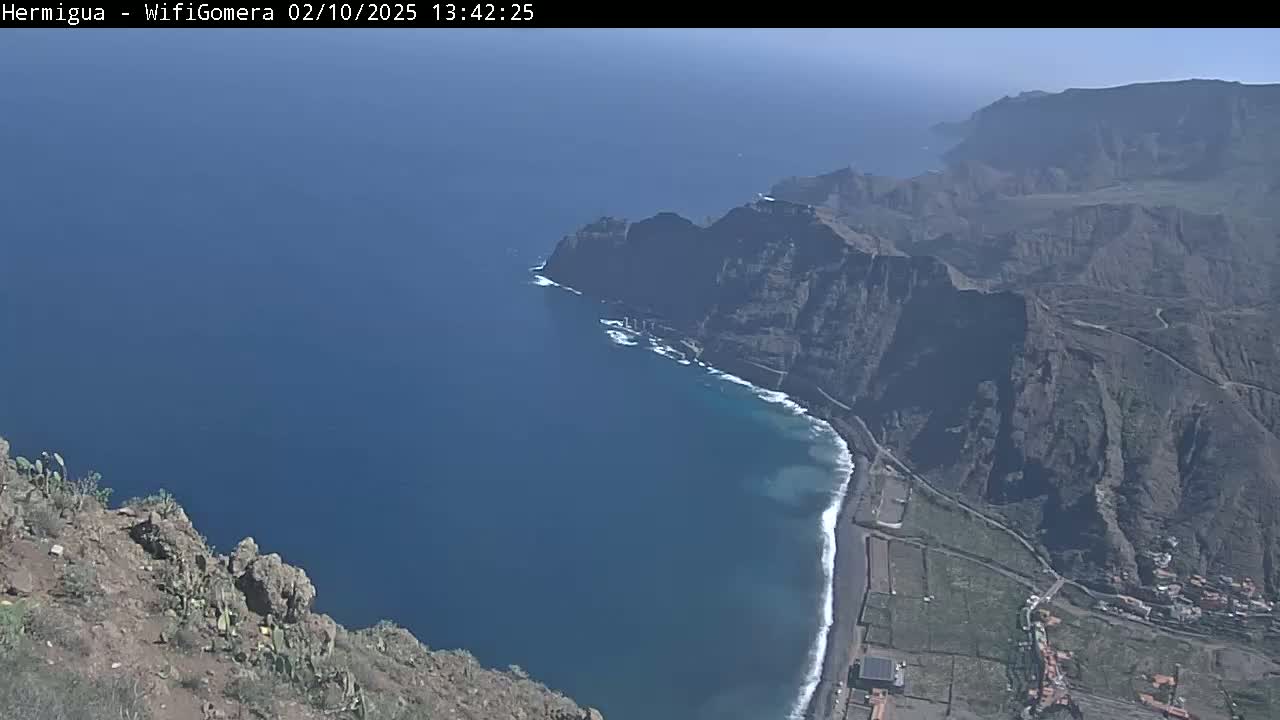 A high-angle view shows a dark-blue ocean meeting a black-sand beach next to dark-brown cliffs and a road under a clear, sunny sky.