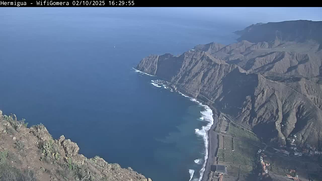 A high-angle view shows a dramatic coastline with dark-colored cliffs meeting a dark-blue ocean where waves crash on a black-sand beach under a clear sky.