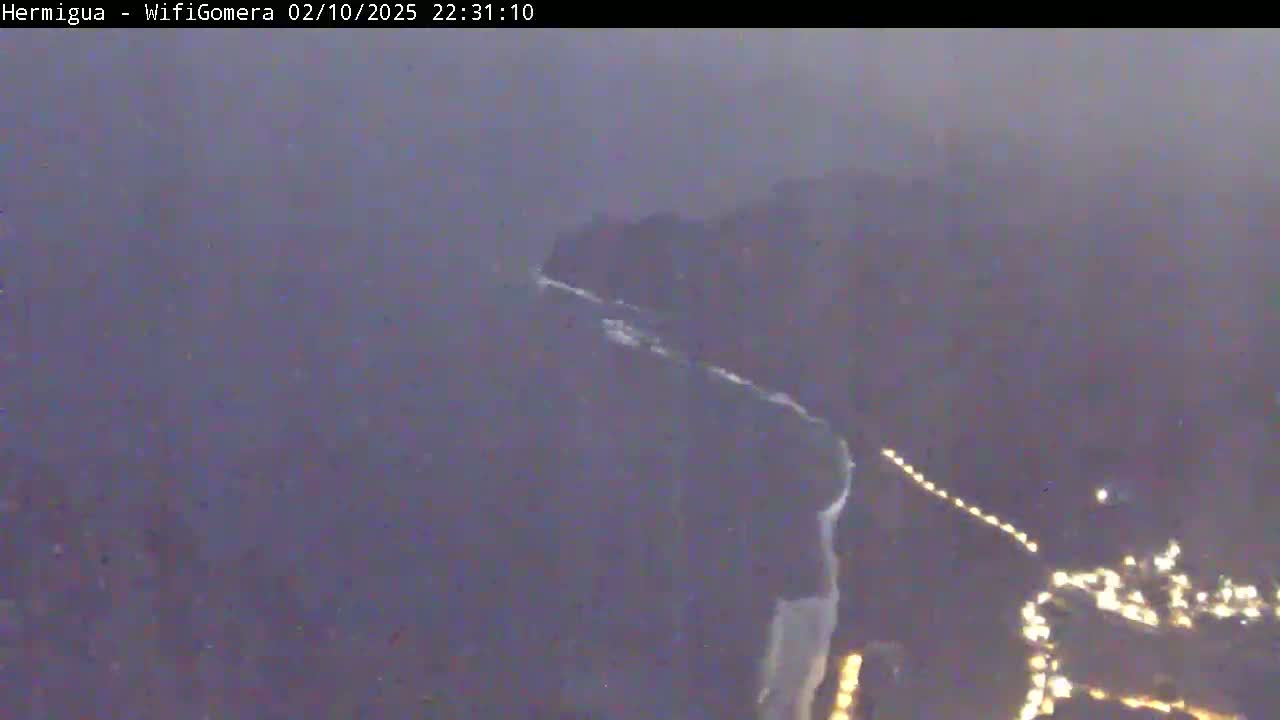 A hazy, dark nighttime view shows a rocky coastline with lights visible along a road in the lower right, and white waves crashing along the shore.
