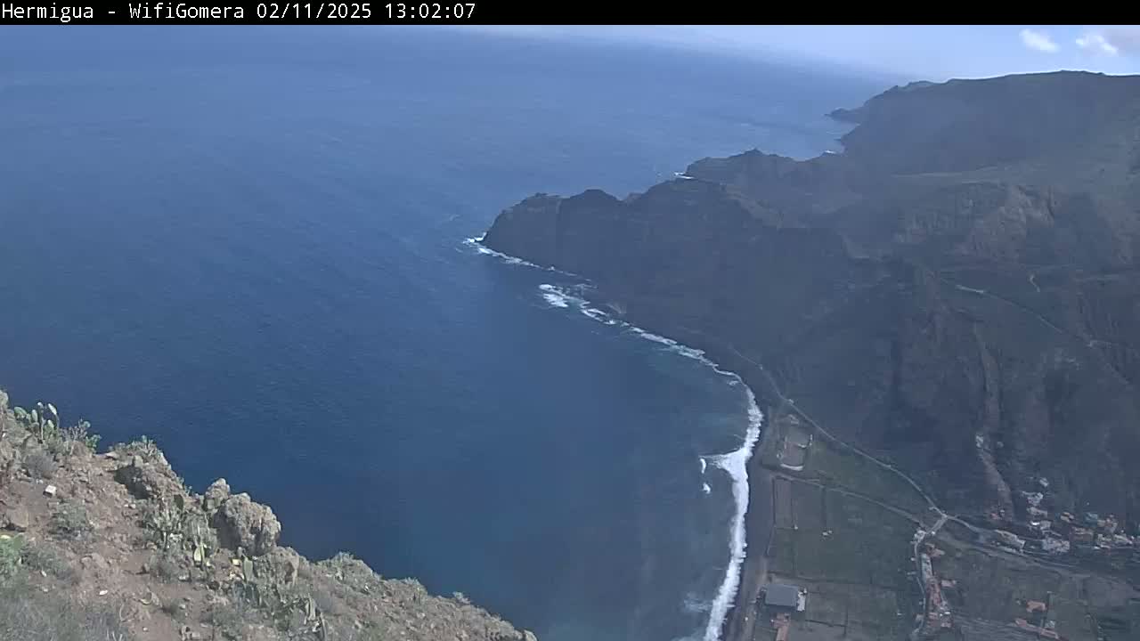 A high-angle view shows a rocky coastline meeting a dark blue ocean under a mostly clear sky.