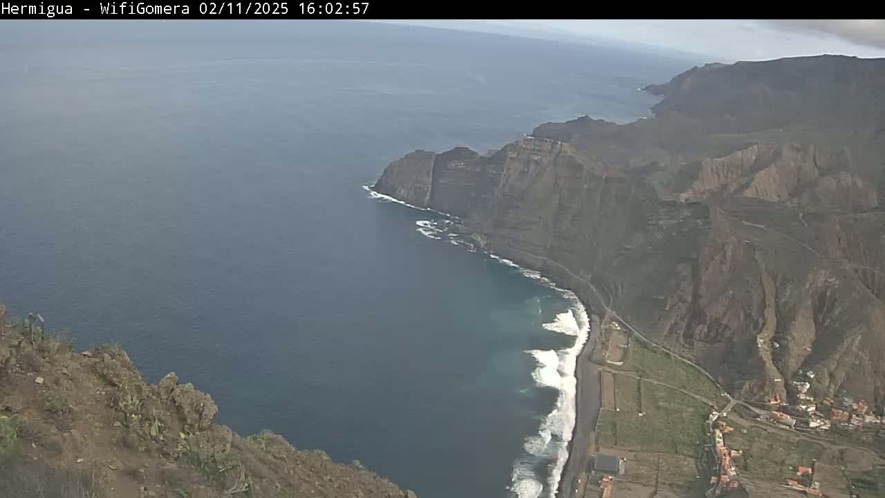A high-angle view shows a rocky coastline meeting a dark blue ocean with small waves crashing against the shore on a partly cloudy day.