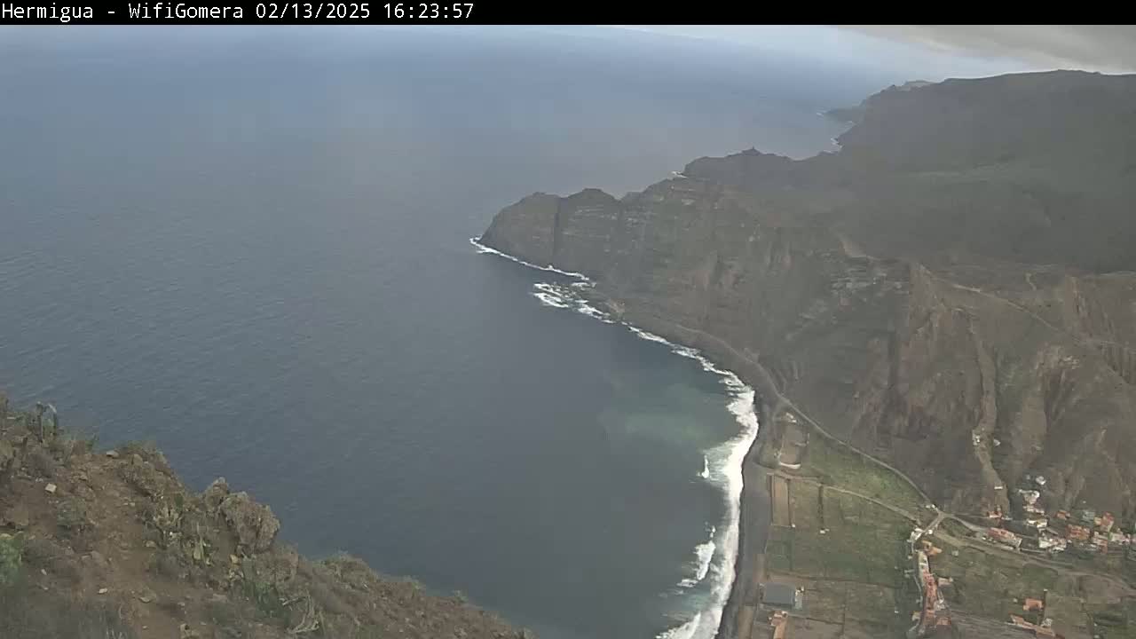 A high-angle view shows a dark-colored ocean meeting a rocky, cliff-lined coast under overcast skies.