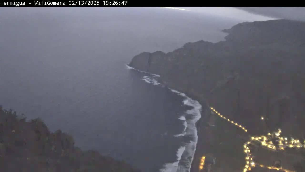 A high-angle, twilight view shows a dark coastline with waves crashing on the shore, a village with lights visible below, and a dark, calm ocean extending into the distance.