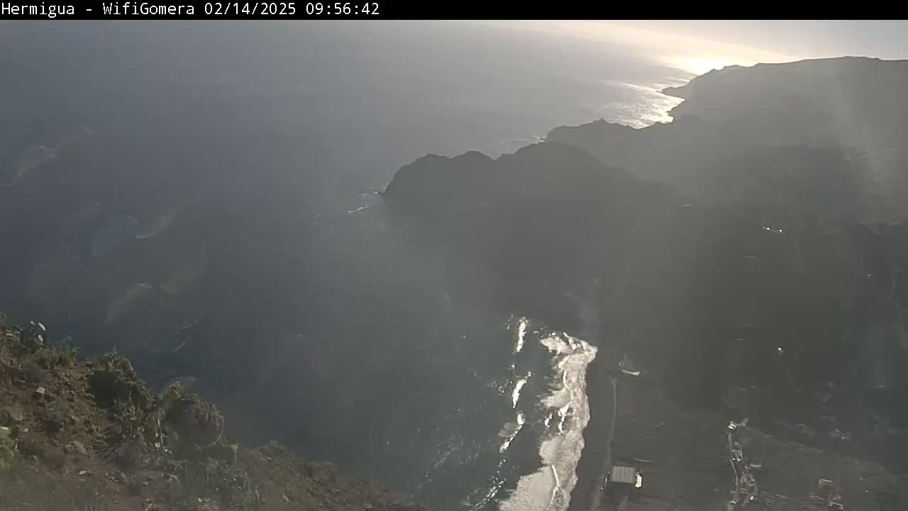 A hazy, sunny view from a high vantage point shows dark volcanic rock formations meeting a dark ocean, with some lighter-colored areas possibly indicating sand or surf along the coast.