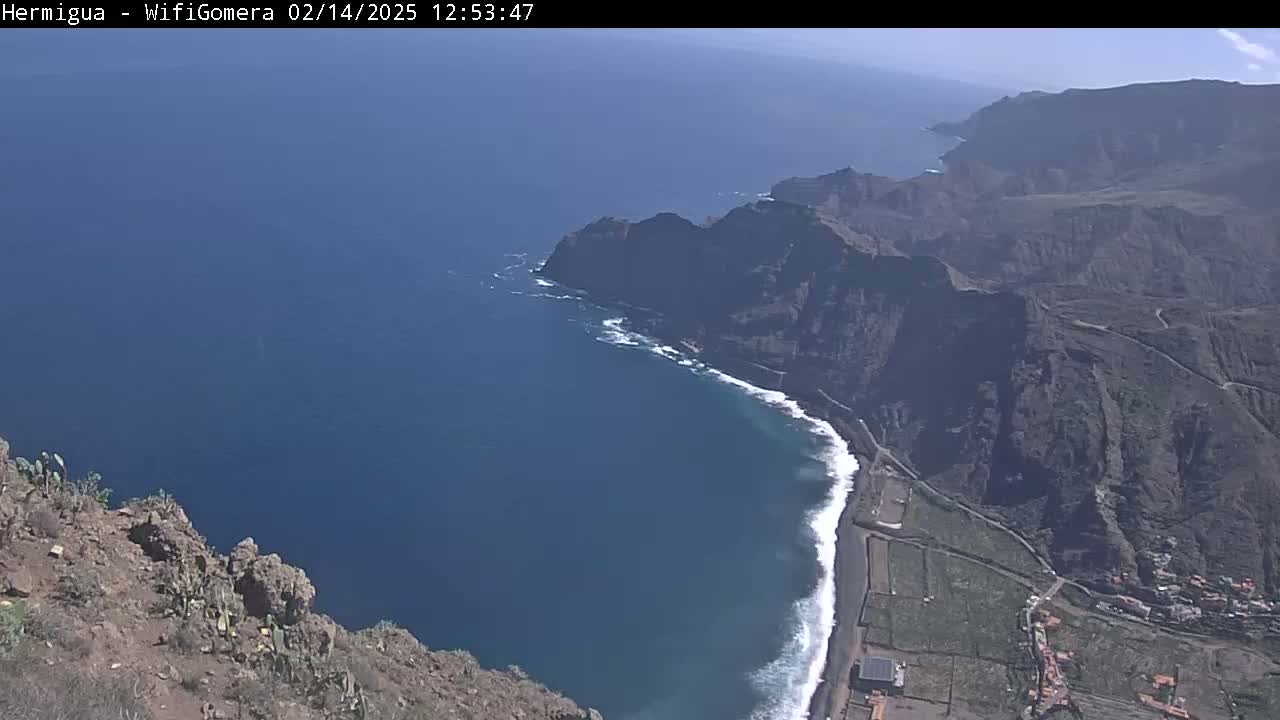 A high-angle view shows a rocky, mountainous coastline meeting a dark-blue ocean with small waves on a sunny day.
