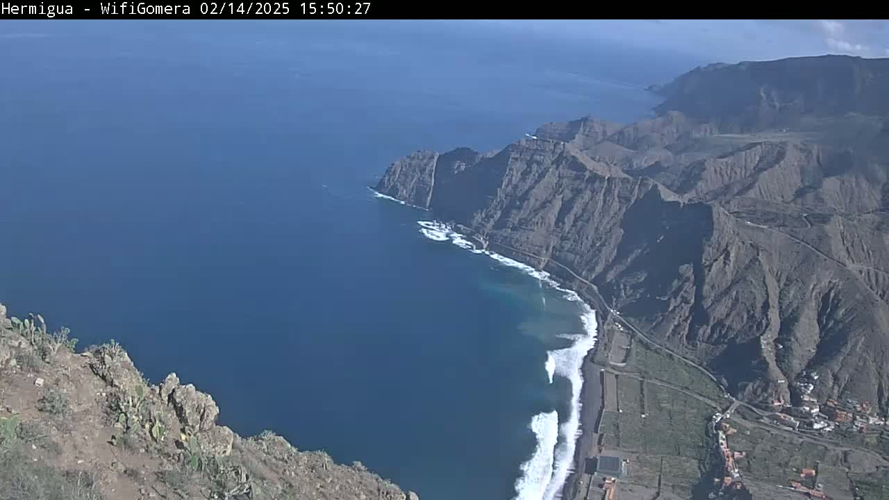 A high-angle view shows a rugged, dark-grey volcanic coastline meeting a deep blue ocean with white-capped waves under a clear sky.