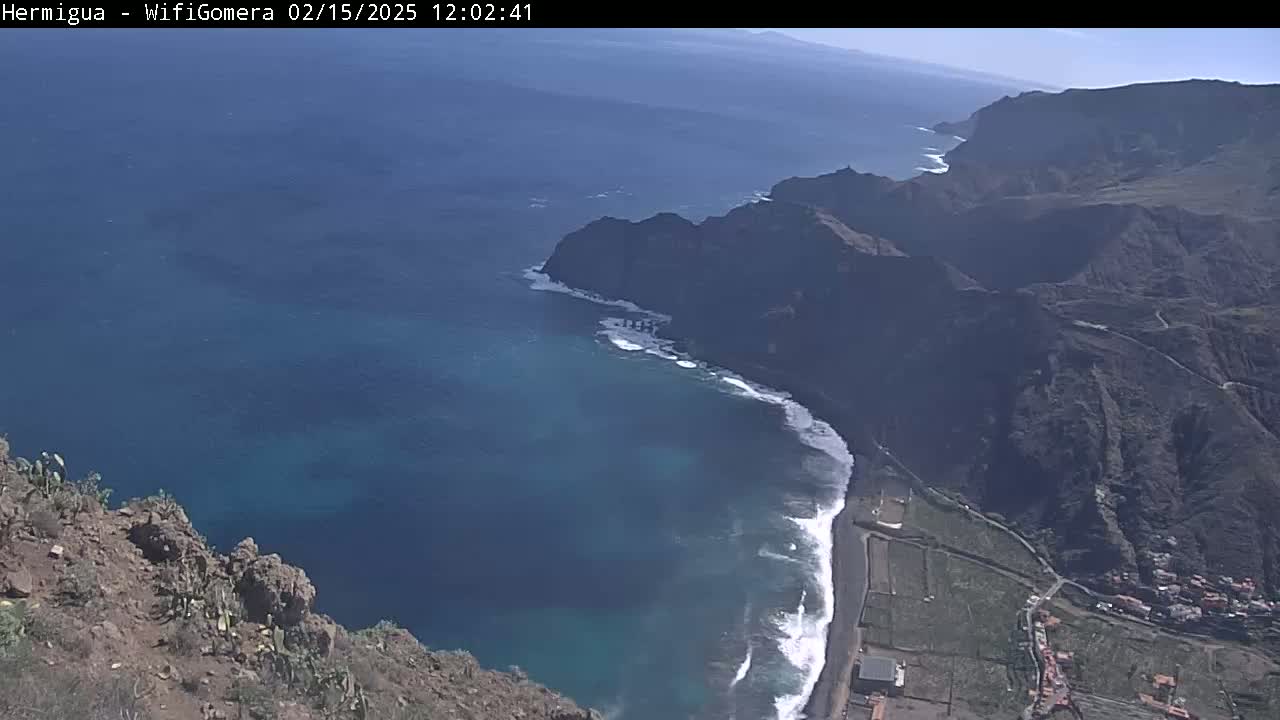 A high-angle view shows a rugged, dark-colored coastline with a small beach where waves break, all under a clear, sunny sky.