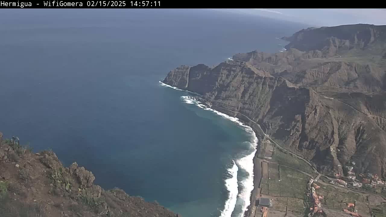 A high-angle view shows a rocky coastline with a dark-blue ocean, white-capped waves crashing against the shore under a clear sky.