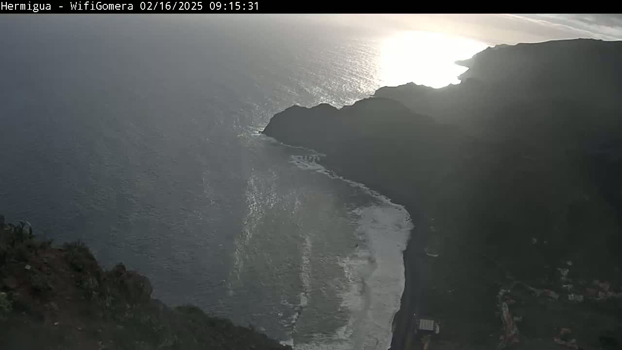 A high-angle, long shot shows a dark, rocky coastline meeting a somewhat choppy ocean under a bright, hazy sun.