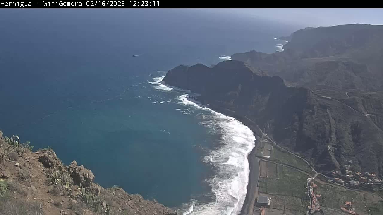 A high-angle view shows a rocky coastline with a dark-blue ocean featuring white-capped waves under a mostly sunny sky.