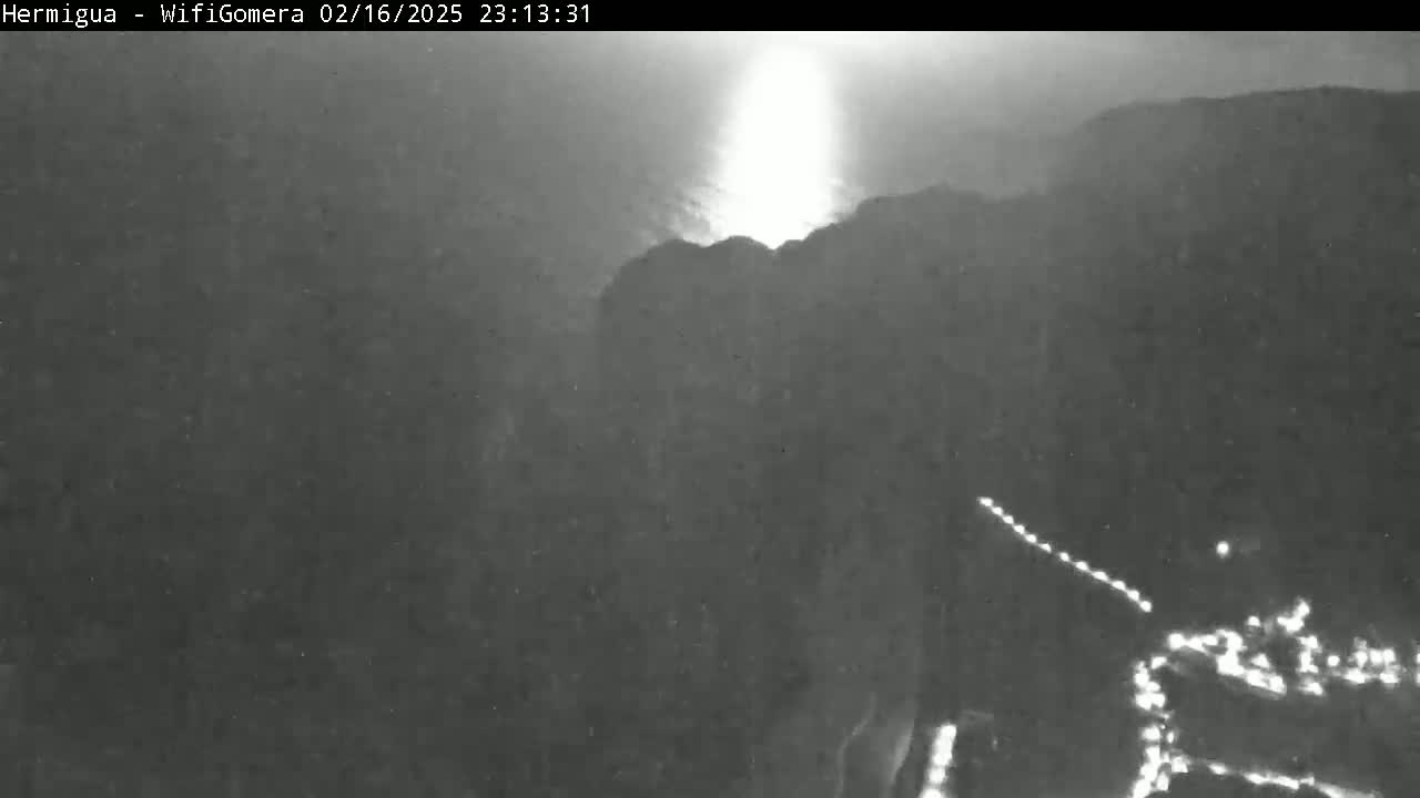 A nighttime, low-light image shows a bright light emanating from behind a dark hill or mountain, with a line of lights visible below.