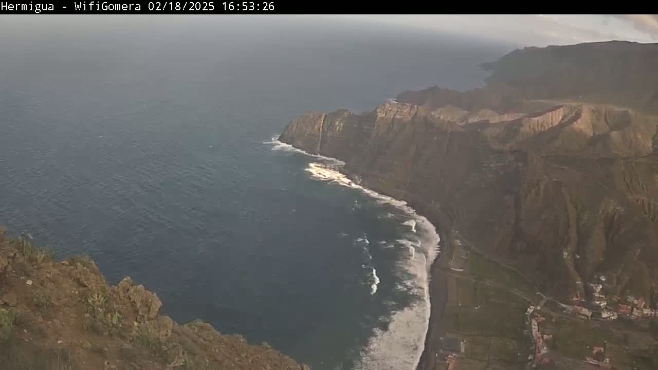 A high-angle view shows a rocky coastline with dark-colored cliffs meeting a dark blue ocean where waves are crashing onto a black sand beach, under slightly hazy conditions.