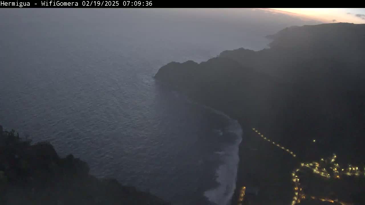 A hazy, pre-dawn view shows a dark coastline meeting a dark ocean with some small waves, a town's lights visible in the lower right corner.