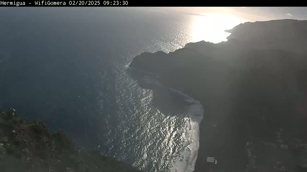 A high-angle, sunny view shows dark cliffs meeting a sparkling ocean under a mostly clear sky.
