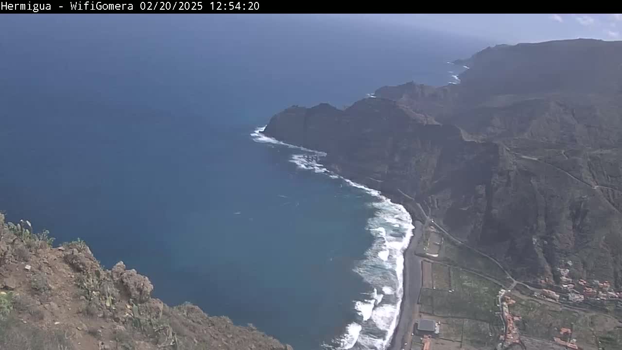 A high-angle view shows a rocky coastline with crashing waves on a mostly sunny day.