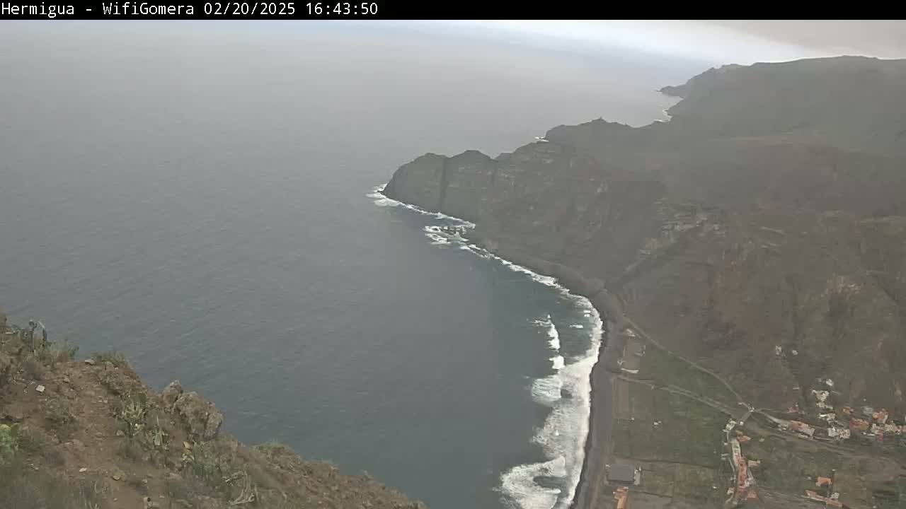 A hazy coastal view shows dark volcanic cliffs meeting a dark-grey ocean with white waves, along a dark-sand beach and sparse green vegetation.