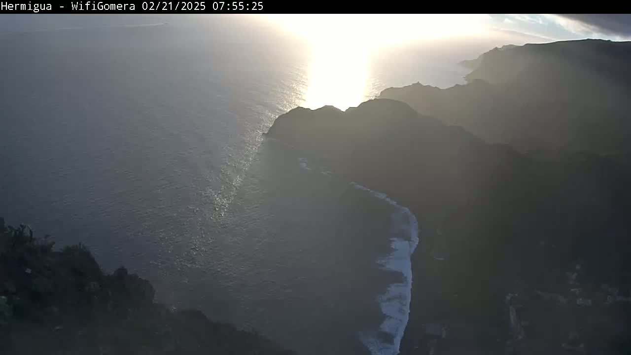 A high-angle view shows a sunlit ocean meeting dark, mountainous coastlines under a partly cloudy sky.