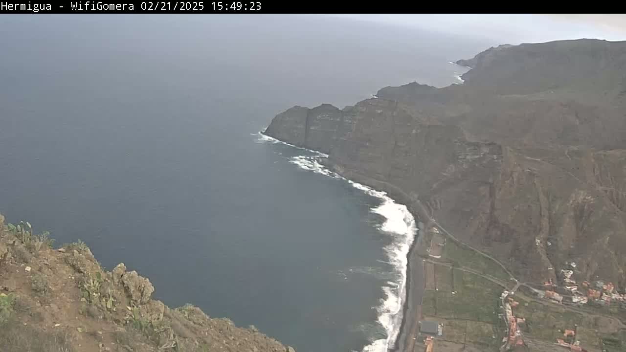 A high-angle view shows dark-colored cliffs meeting a dark-blue ocean with white-capped waves under an overcast sky.