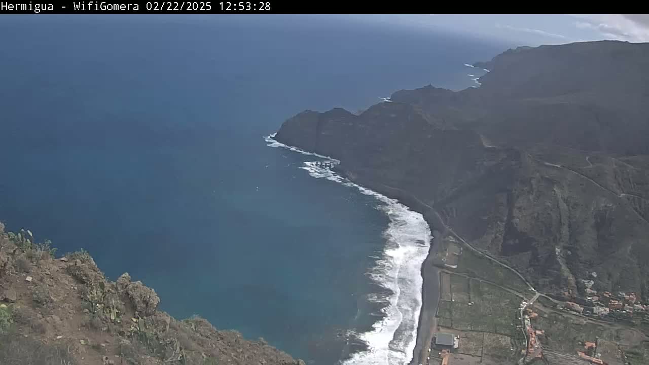 A high-angle view shows a dark-blue ocean meeting a black-sand beach that curves along a rocky, dark-brown coastline on a partly sunny day.