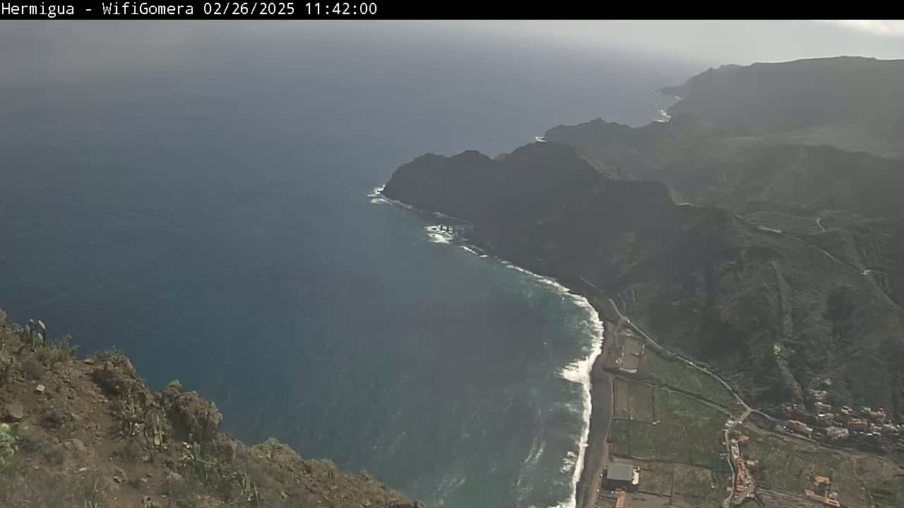 A high-angle, hazy view shows a dark-colored coastline with a dark blue ocean, meeting a mountainous landscape.