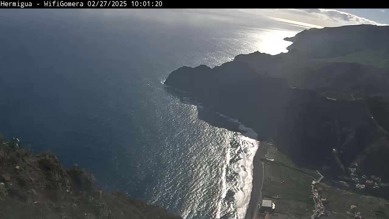 A high-angle view shows a rocky coastline meeting a bright, wavy ocean under a mostly sunny sky.