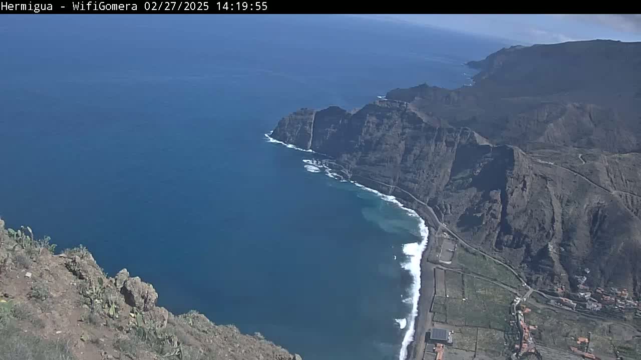 A high-angle view shows a rugged, dark-colored coastline meeting a deep blue ocean under sunny skies, with waves breaking along a dark-sand beach.