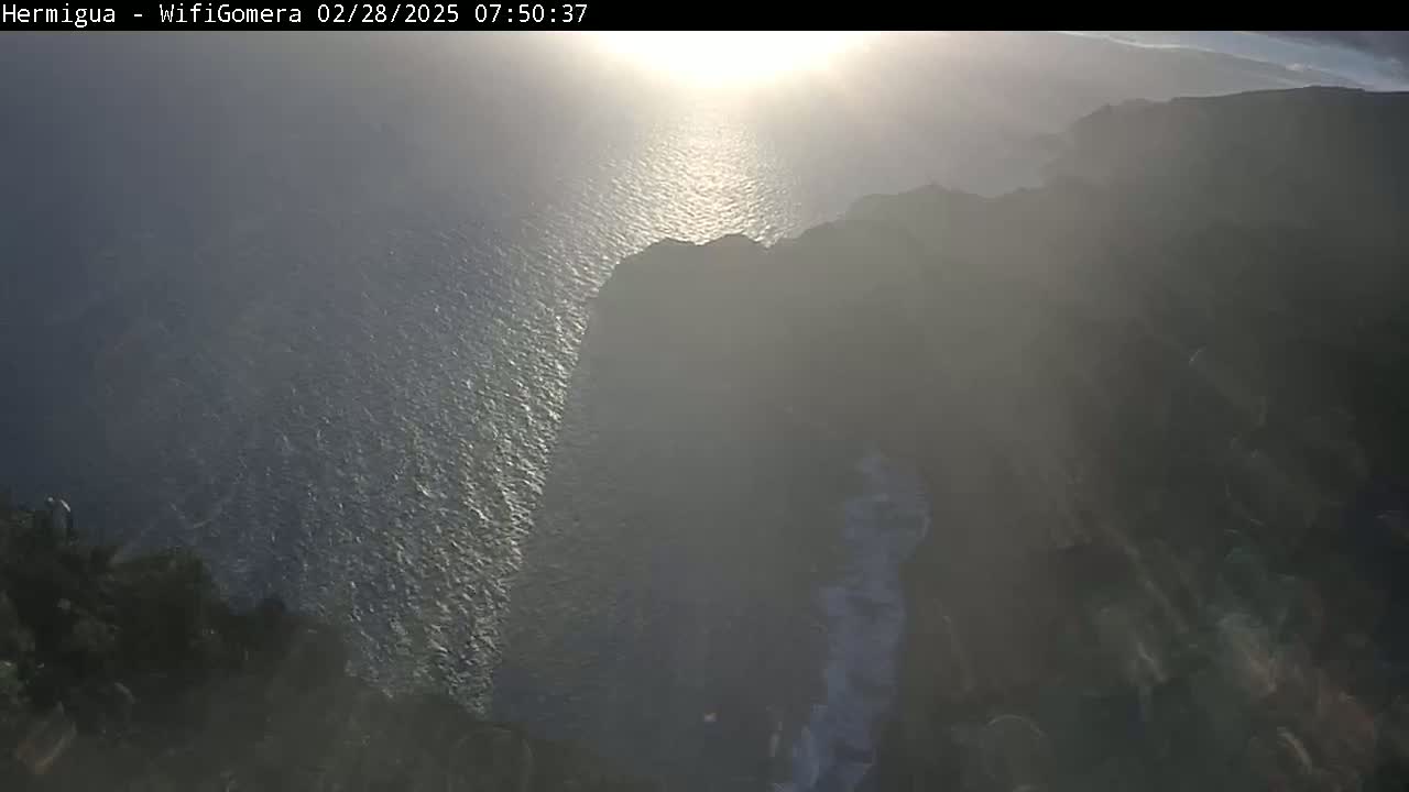 A hazy sunrise illuminates a dark cliff overlooking a sparkling ocean.