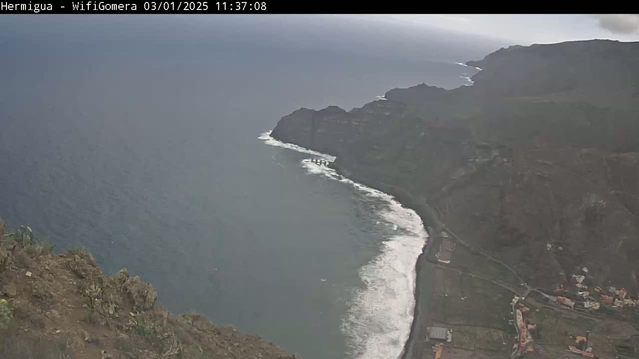 A high-angle view shows a rocky coastline with dark volcanic cliffs meeting a dark-blue ocean with white-capped waves under an overcast sky.