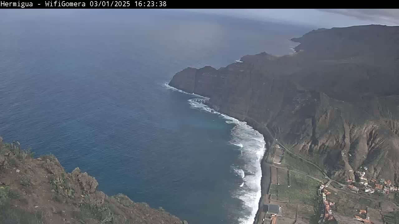 A high-angle view shows dark, rocky cliffs meeting a dark-blue ocean with white waves along a shoreline, under mostly cloudy skies.