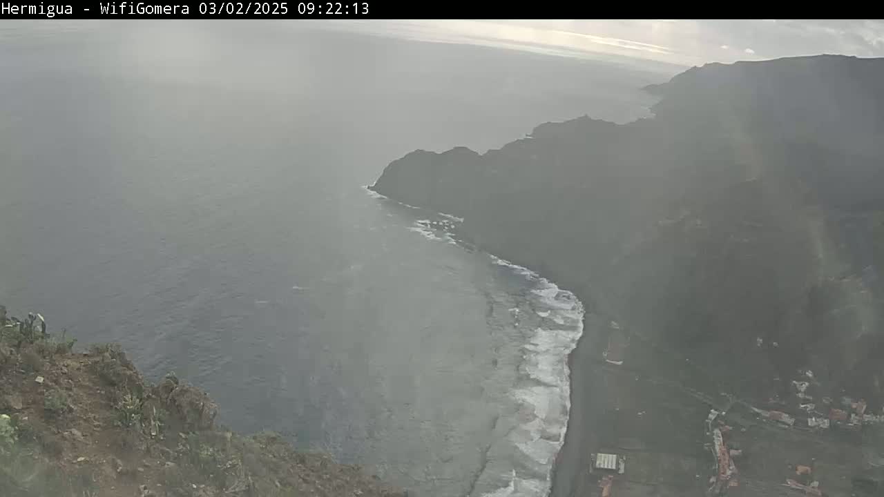 A hazy view shows a rocky cliff overlooking a dark ocean with waves crashing on a beach below, a small town visible at the base of the cliff.