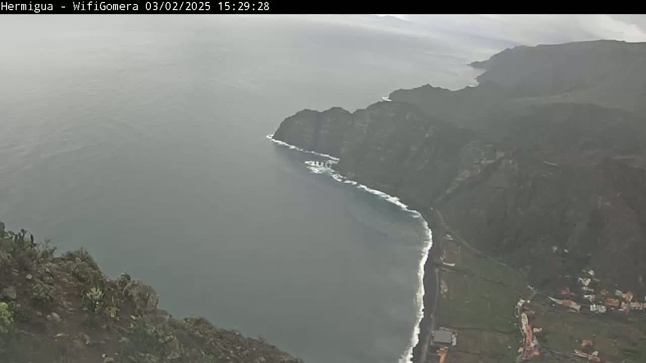 A hazy, overcast view shows a rocky coastline meeting a dark gray sea with small waves, a dark cliff face rising from the ocean to a green hillside.