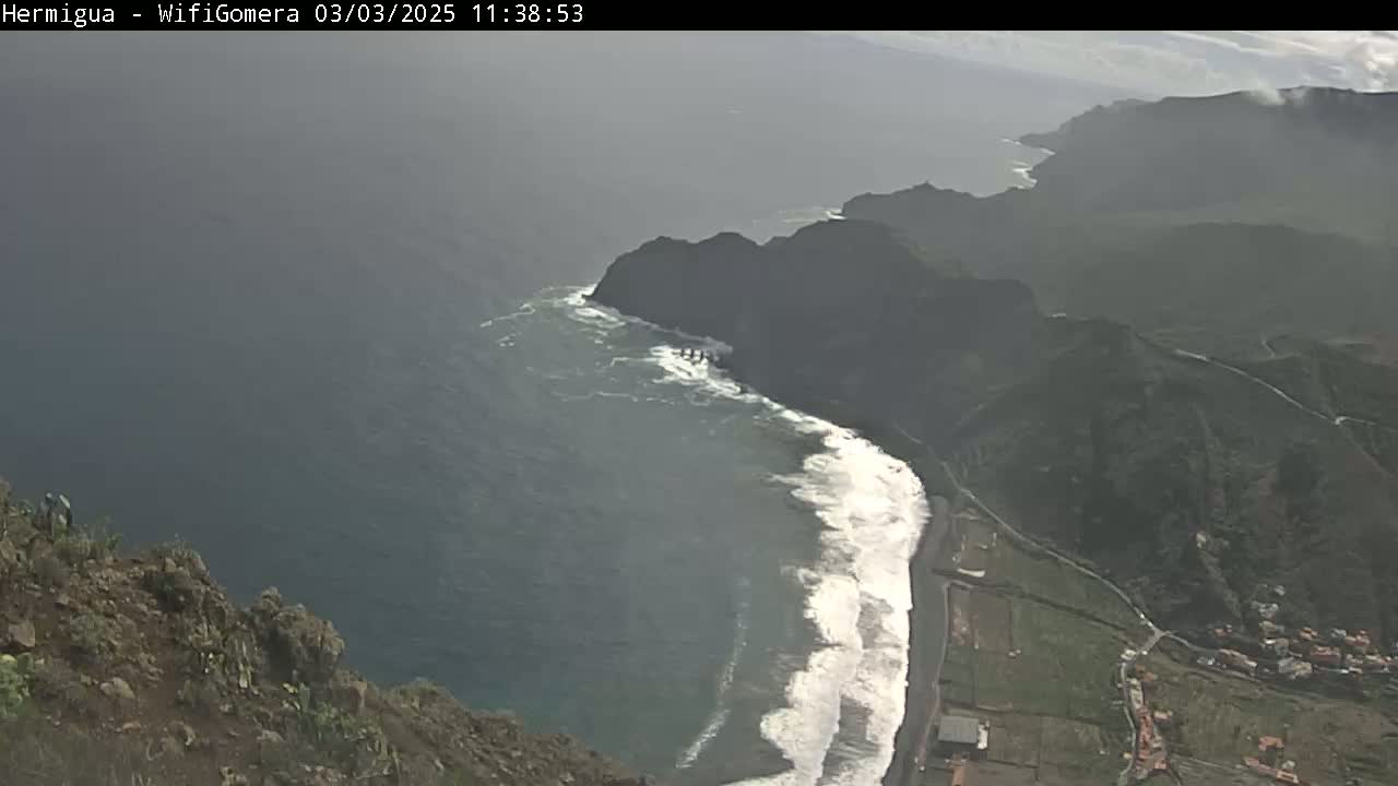 A hazy, high-angle view shows a dark, rocky coastline with crashing waves meeting a dark-colored ocean.