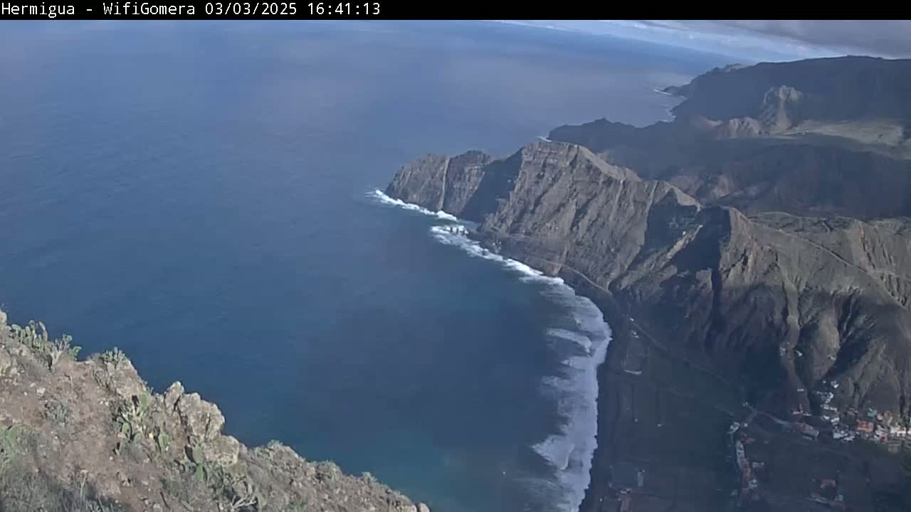 A high-angle view shows a rocky coastline with waves crashing against it, under a mostly clear sky.