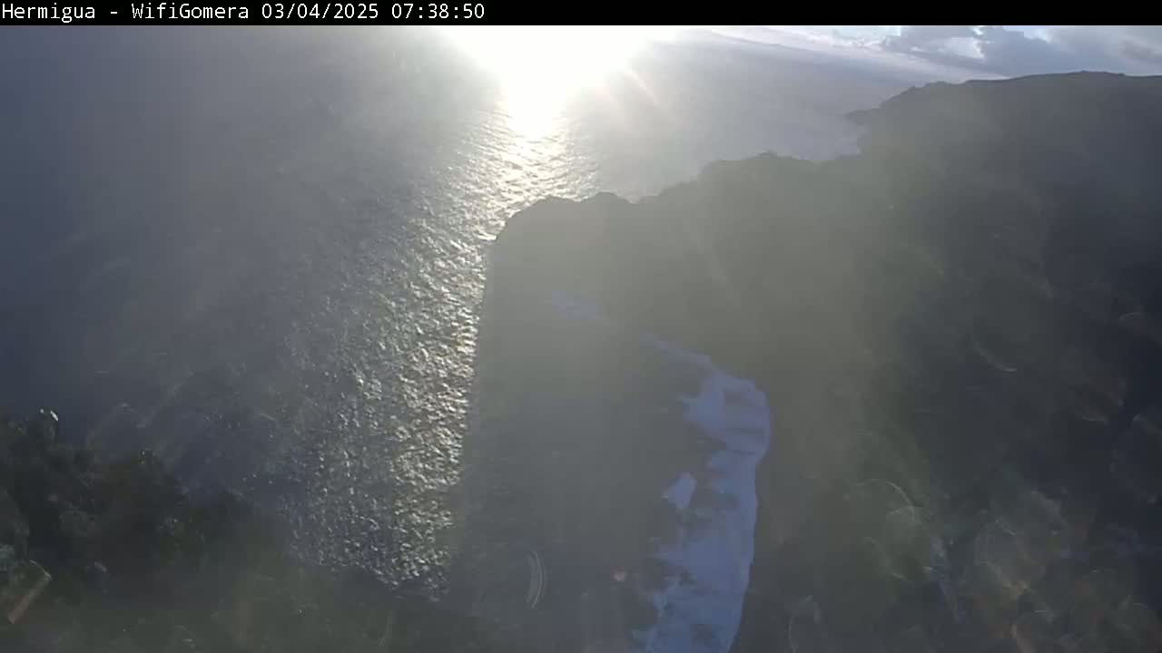 A hazy sunrise illuminates a dark, rocky coastline meeting a bright, wavy ocean.