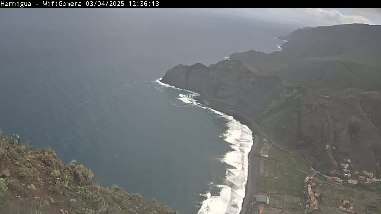 A high-angle, overcast view shows a dark-colored beach meeting dark cliffs that rise to a mountainous landscape overlooking a dark-blue ocean with white-capped waves.