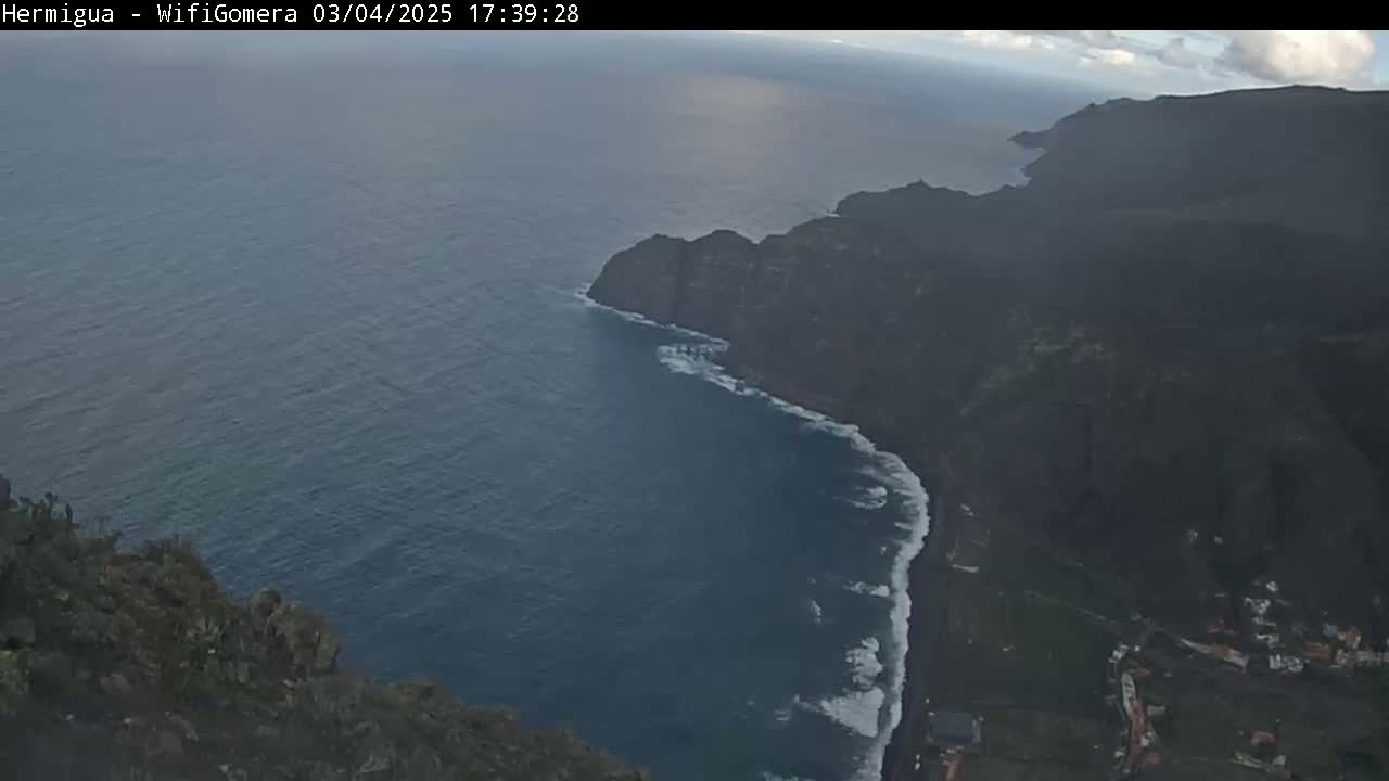 A high-angle view shows a dark, rocky coastline meeting a deep blue ocean with white-capped waves under a partly cloudy sky.