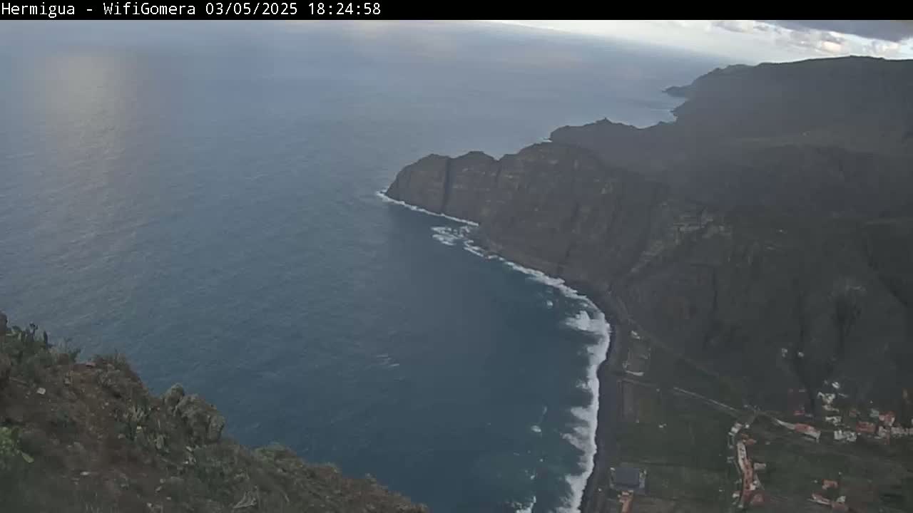 A high-angle view shows dark, rocky cliffs meeting a dark blue ocean with white waves crashing against the shore under an overcast sky.