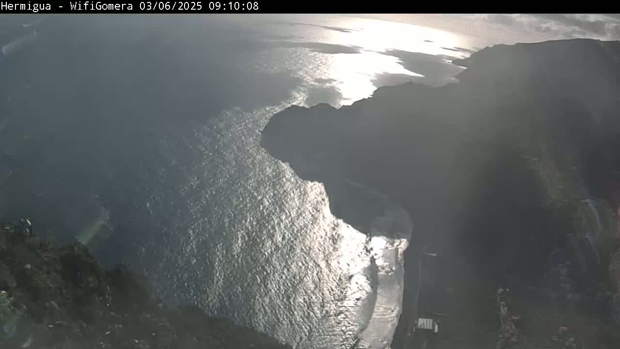 A high-angle view shows a rocky coastline meeting a bright, sunlit ocean under a mostly clear sky.