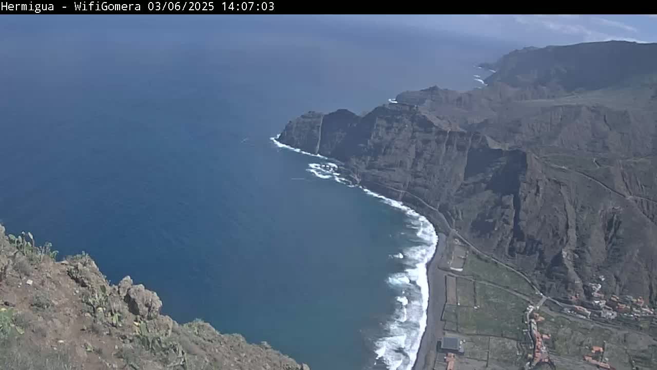 A high-angle view shows a rocky coastline with crashing waves on a sunny day, overlooking a dark-blue ocean and a small town nestled in a valley.