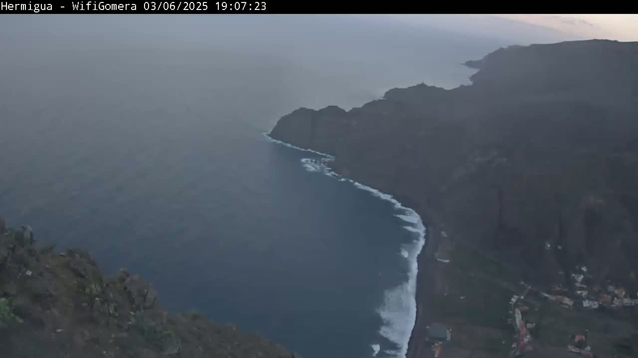 A hazy, high-angle view shows a dark-colored, rocky coastline meeting a dark-blue ocean with white-capped waves.