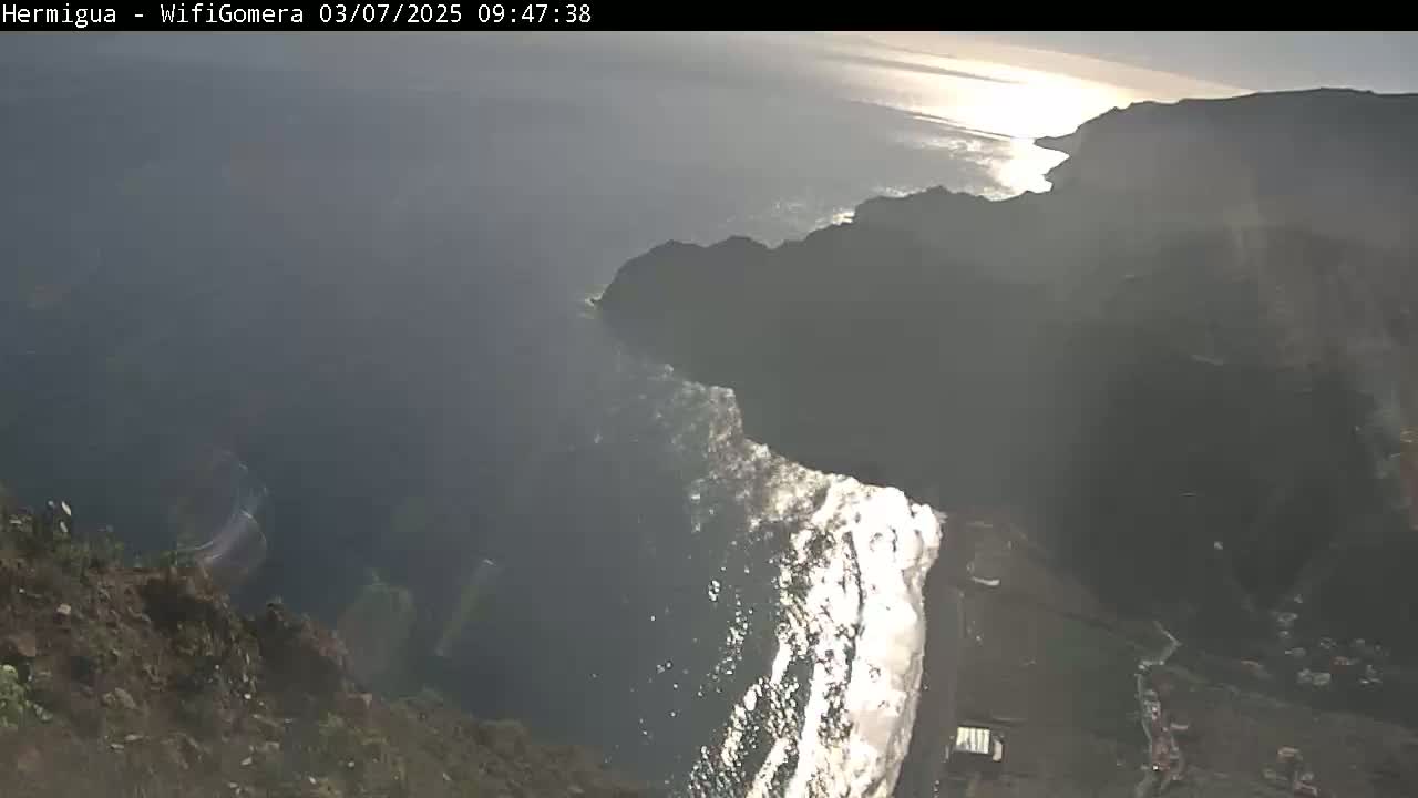 A high-angle, long shot shows a rocky cliff overlooking a dark-grey ocean with white waves crashing onto a shoreline below, under a partly sunny sky.