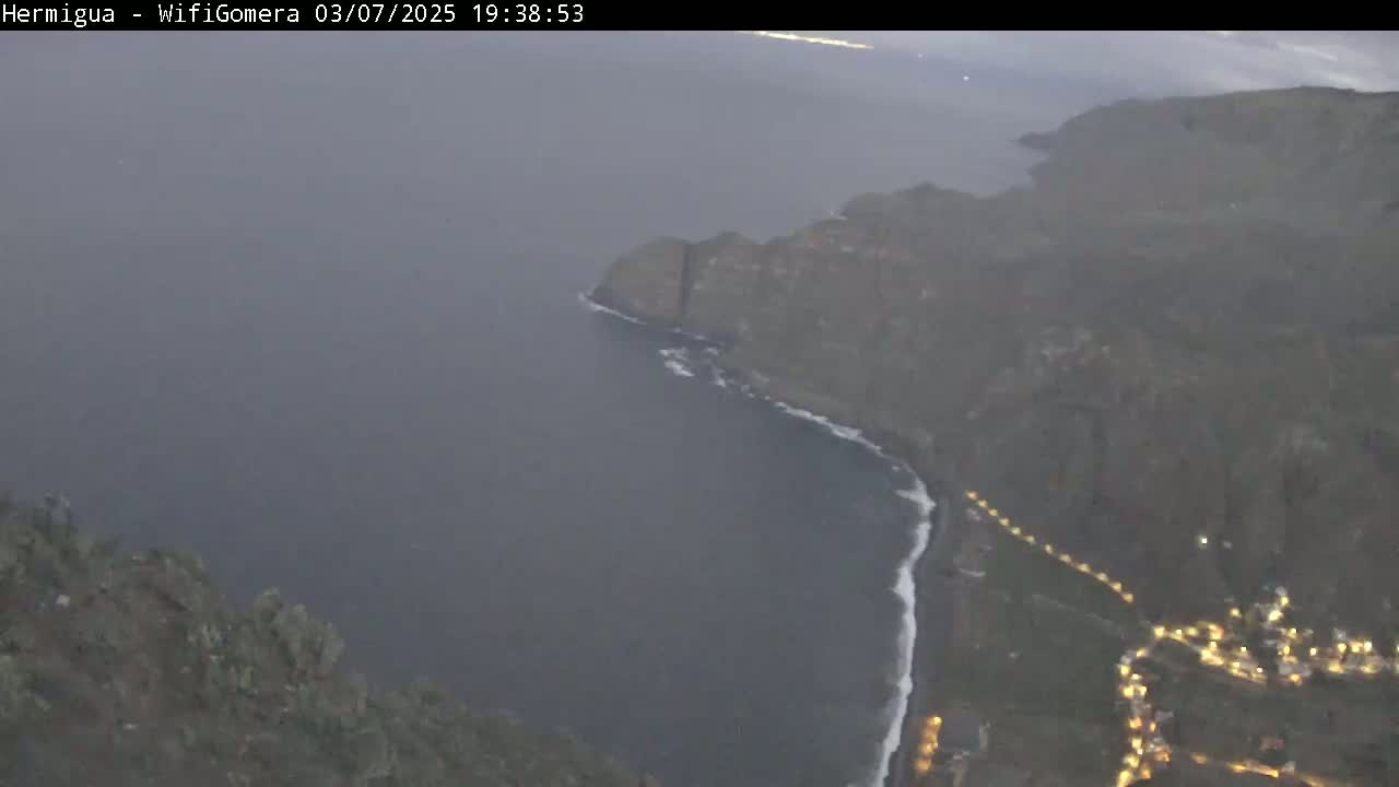 A hazy, high-angle view shows a dark-colored coastline with a village and road at the bottom, meeting a dark ocean; the sky is overcast.