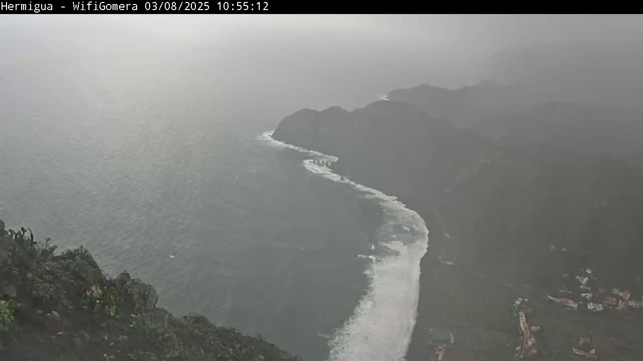 A rain-swept coastal scene shows a dark, rocky shoreline with white waves crashing against it, viewed from a high vantage point.